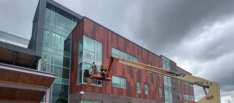 A professional window cleaner in a safety harness operating from an orange boom lift to access the exterior glass of a modern commercial building under an overcast sky.