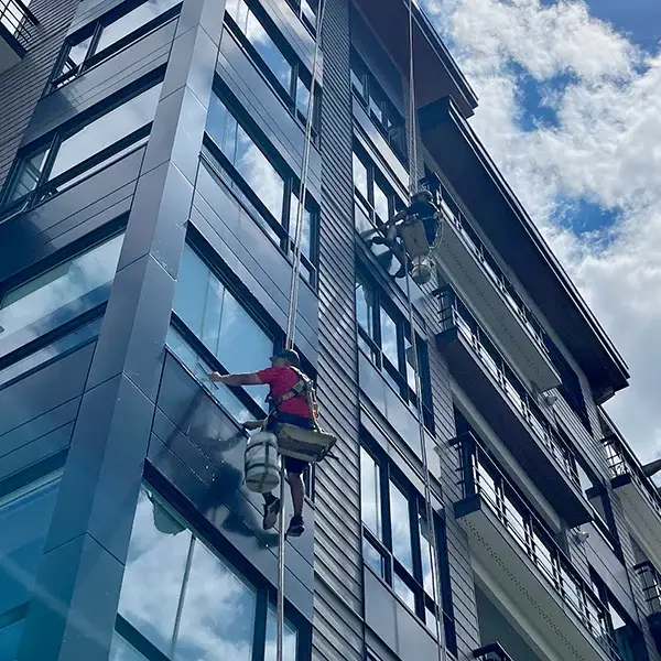 A low-angle view of professional technicians performing Modern mid-rise residential Window Services by rappelling down the exterior of a contemporary glass and metal building under a bright, cloudy sky.