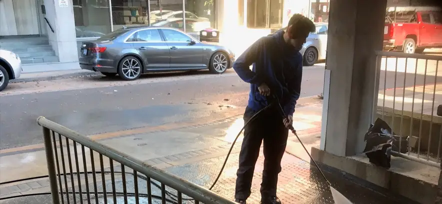 A young man in a blue hoodie and black pants is pressure washing a concrete floor inside a parking garage or covered walkway. He is wearing red ear protection and focused on the task. The high-pressure stream of water is actively stripping away dark grime, revealing the lighter, clean concrete underneath. In the background, a city street is visible with several parked cars, including a grey sedan and a red pickup truck, under bright daylight.