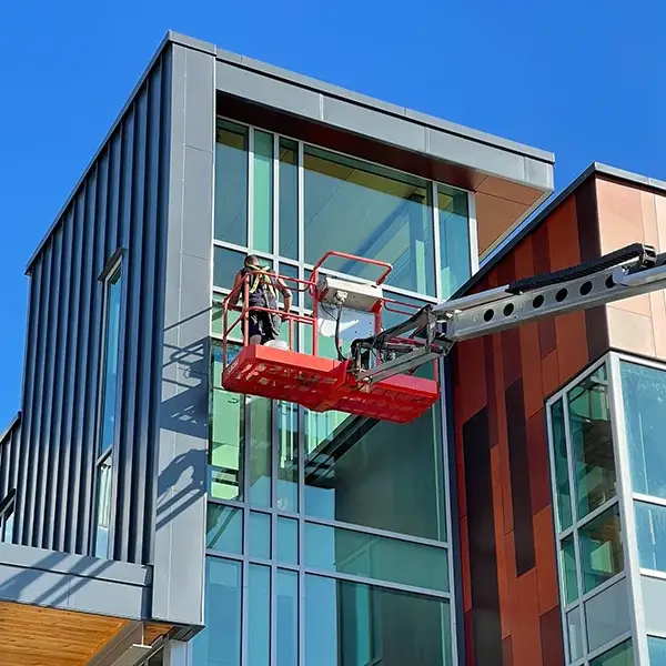 A professional high-altitude service in action. A window cleaner uses a scissor lift to access the windows of a contemporary commercial building, isolated against a clear blue sky for web design use.