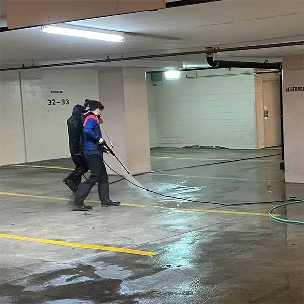 There are two individuals involved in the cleaning process. One person in the foreground, wearing a blue and red jacket, black pants, and waterproof boots, is actively operating a long power washing wand. A second person, dressed in dark clothing, is walking behind them, likely managing the hose or assisting with the equipment.