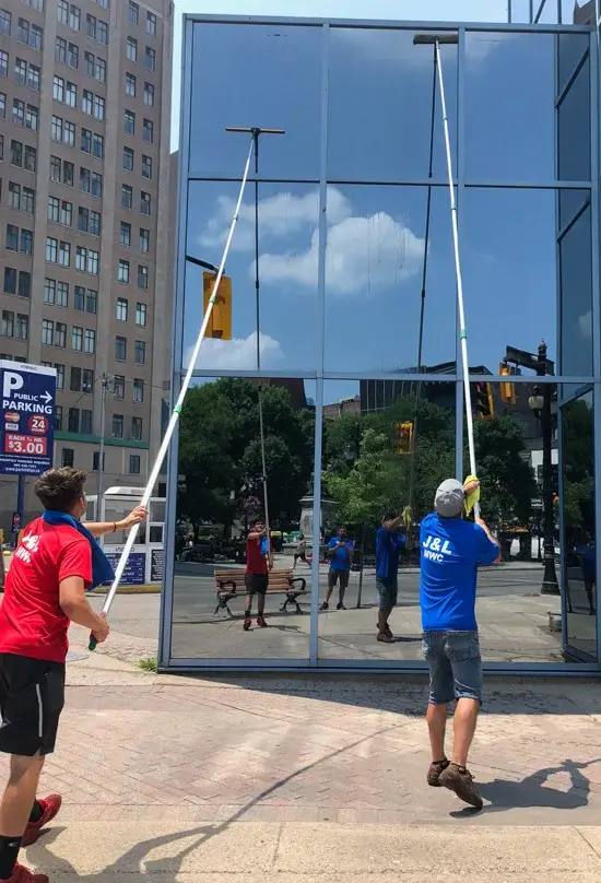 An image of two professional technicians performing High Rise Window Cleaning on a large glass building using long telescopic extension poles. One worker in a red shirt and another in a blue shirt stand on a brick sidewalk, reaching high up the facade. The glass reflects a city street, a tall brick building, and a blue sky with light clouds.