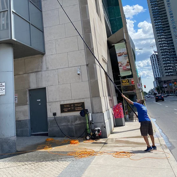 A worker in a blue t-shirt and plaid shorts stands on a city sidewalk, using an exceptionally long, telescoping water-fed pole to clean the exterior windows of a large stone building. On the ground beside him is a portable water filtration system and several tangled coils of orange hosing. The scene is set in a bright, sunny downtown area with a tall skyscraper and city traffic visible in the background.